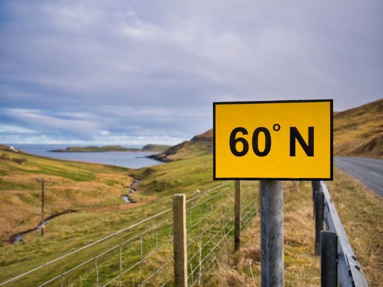 A rectangular, yellow sign with black figures on the A970 on Mainland, Shetland, UK marks the 60 degree North line of latitude. The water of Channer Wick appears on the left.
