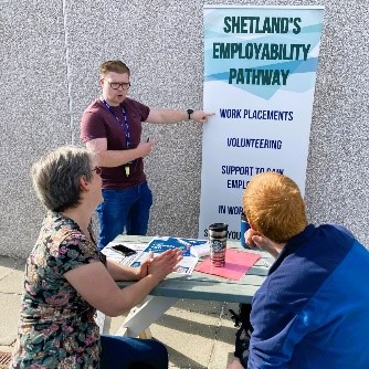 People sitting outside at a table with one person standing and pointing at a banner with the words, 'Shetland's employability pathway' at the top.