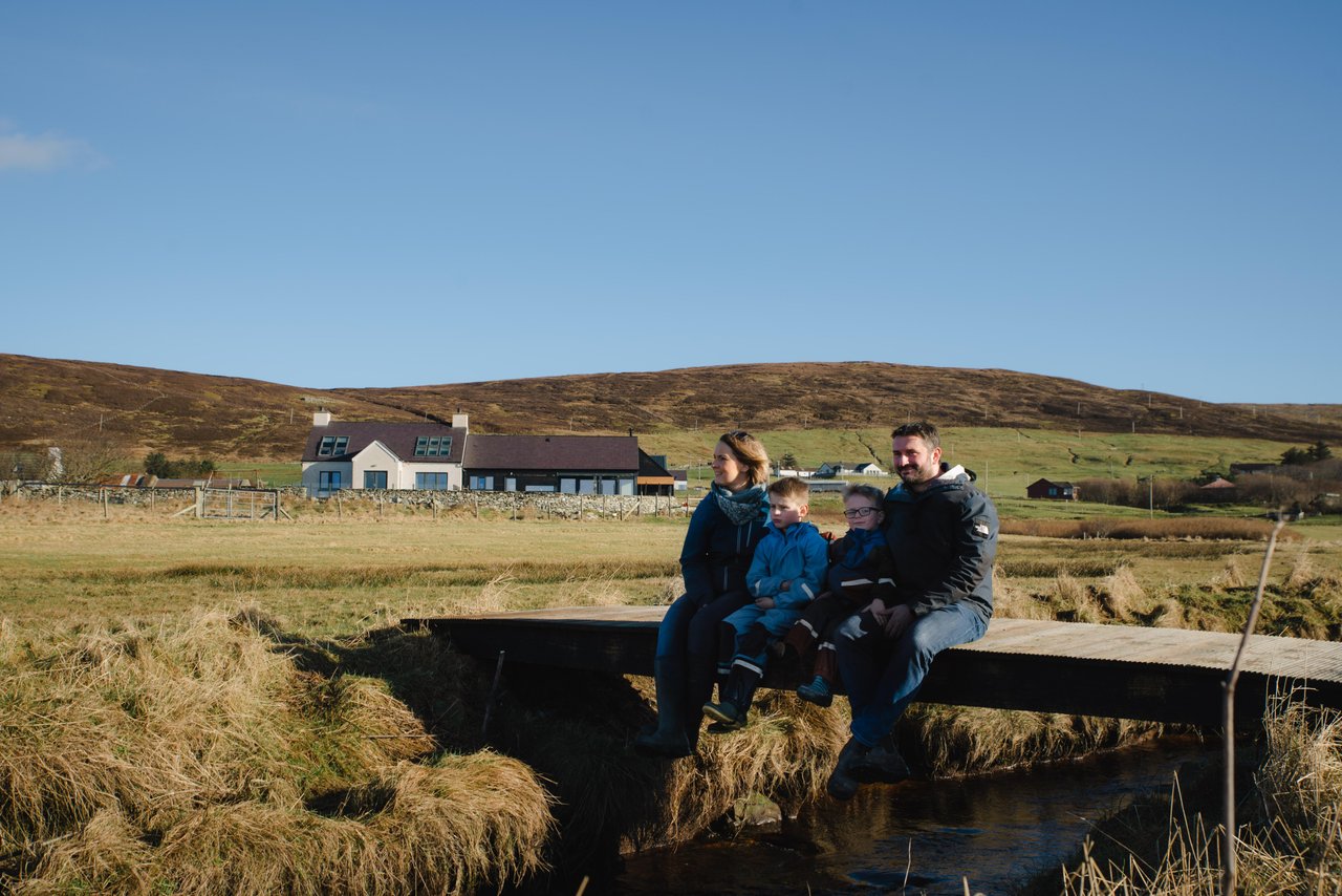 A family sitting together outdoors with their home and the Shetland landscape behind them.