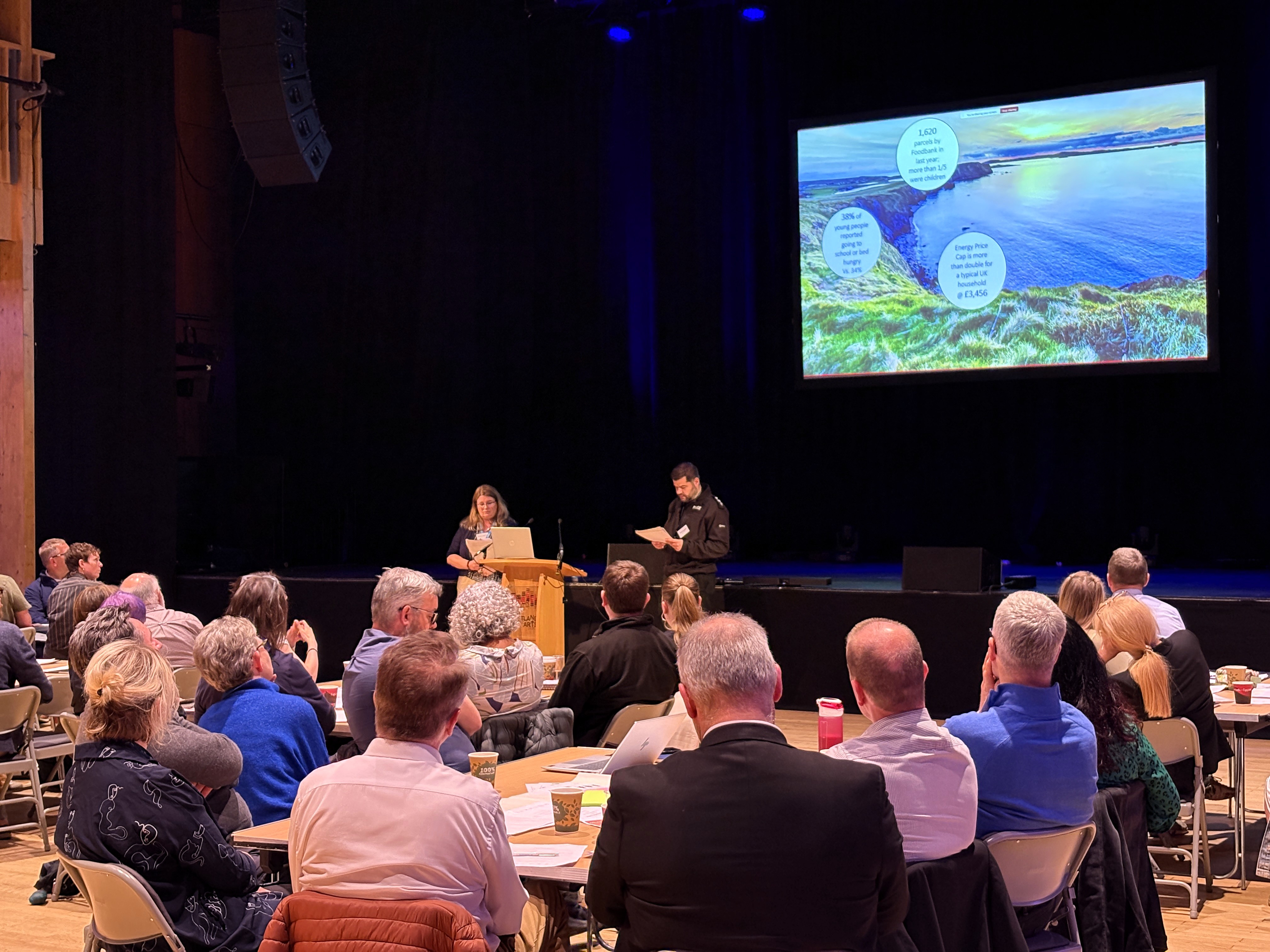 A group of people viewing a presentation with two speakers facing them.