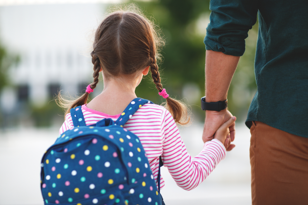 Young girl with a blue backpack, pink top, and pigtails. She is holding hands with an adult, only the hand and part of the body is on display.
